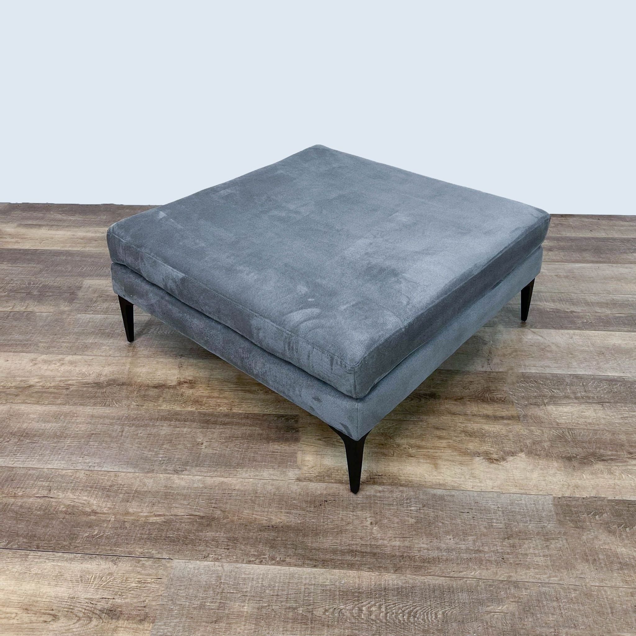 Bauhaus Furniture's Scandinavian-inspired ottoman in gray with a square form and dark-finished metal legs, set against a wood floor.