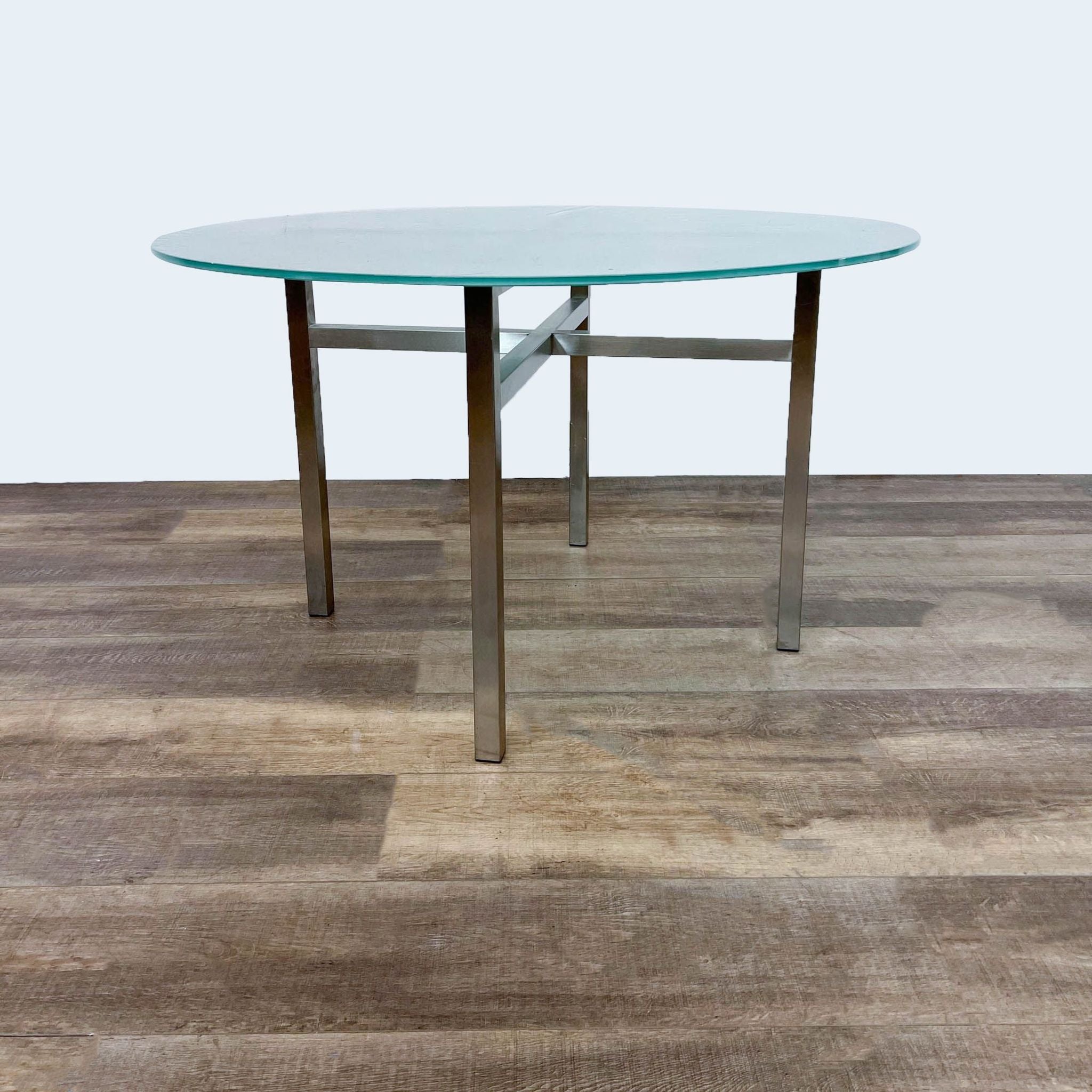 Room & Board Benson dining table with gold finish metal cross brace base and round frosted tempered glass top on a wooden floor.