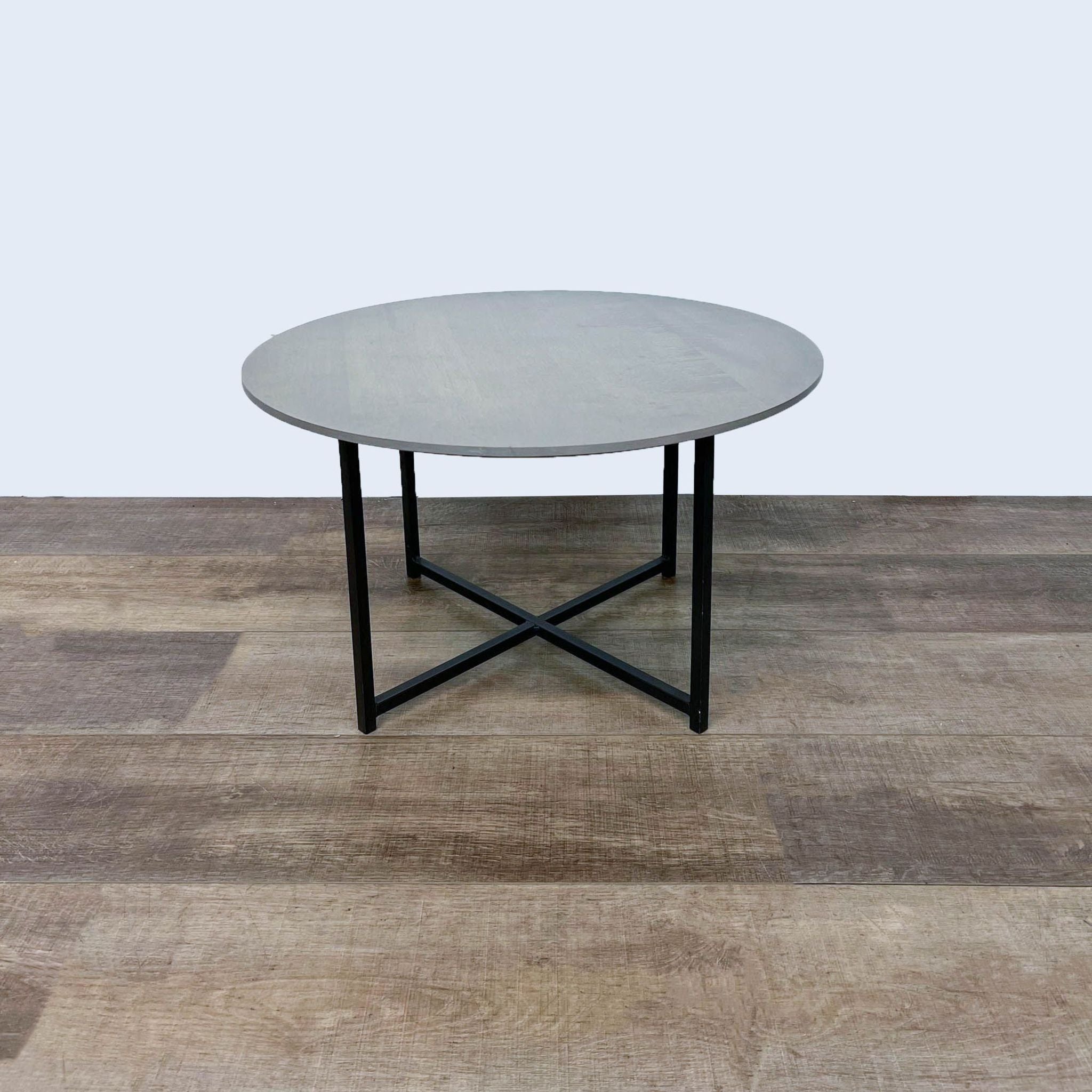 Room & Board coffee table featuring a circular shell-stained maple top and cross steel base, set against a wooden floor backdrop.