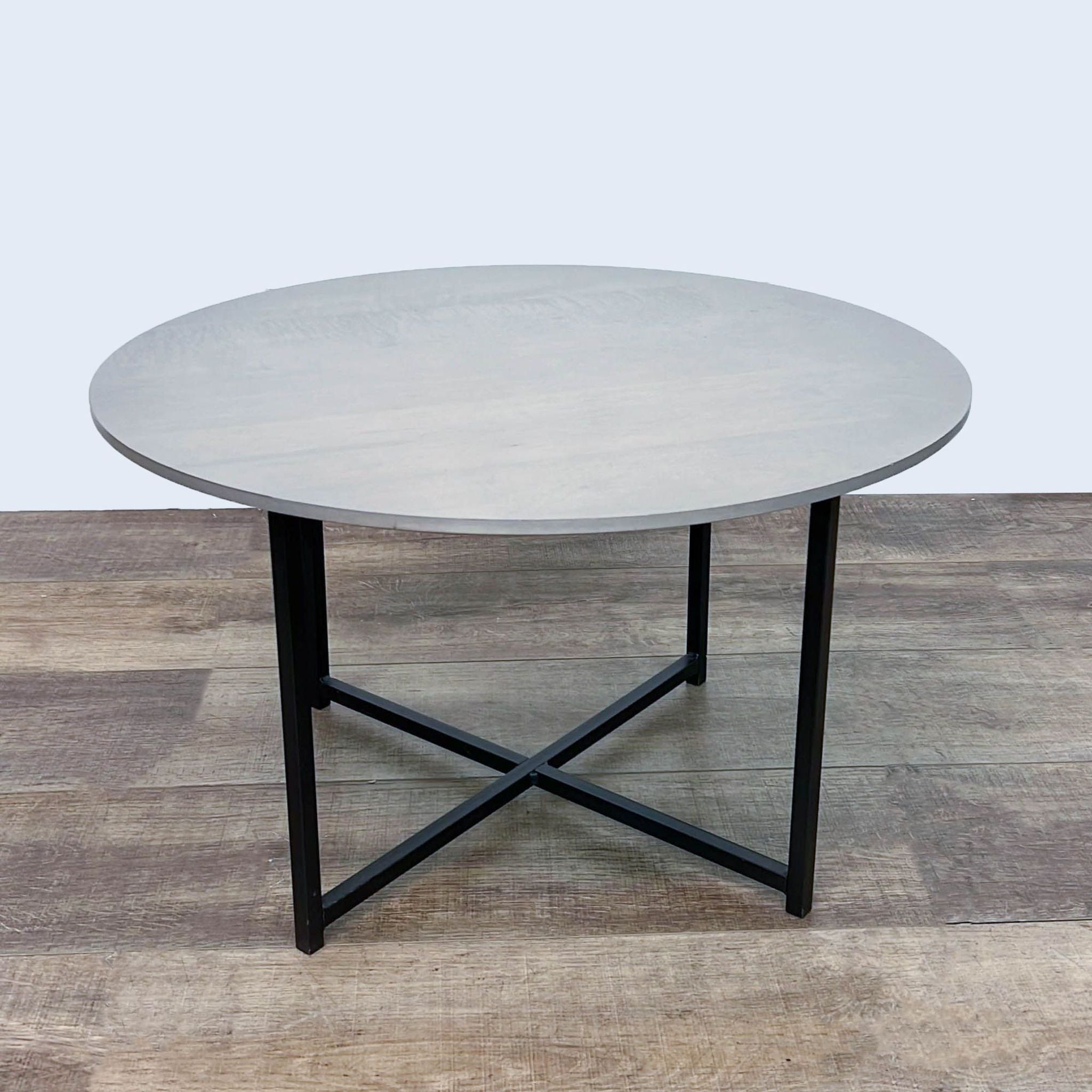 Round coffee table by Room & Board with maple top in shell stain and X-shaped natural steel base on a wooden floor.
