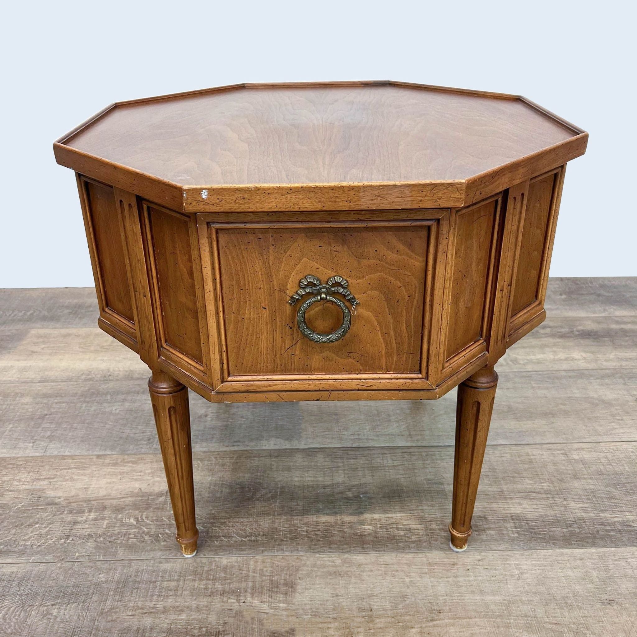 Hexagonal wooden end table by Reperch with a closed drawer and ornate metal pull, standing on tapered legs.