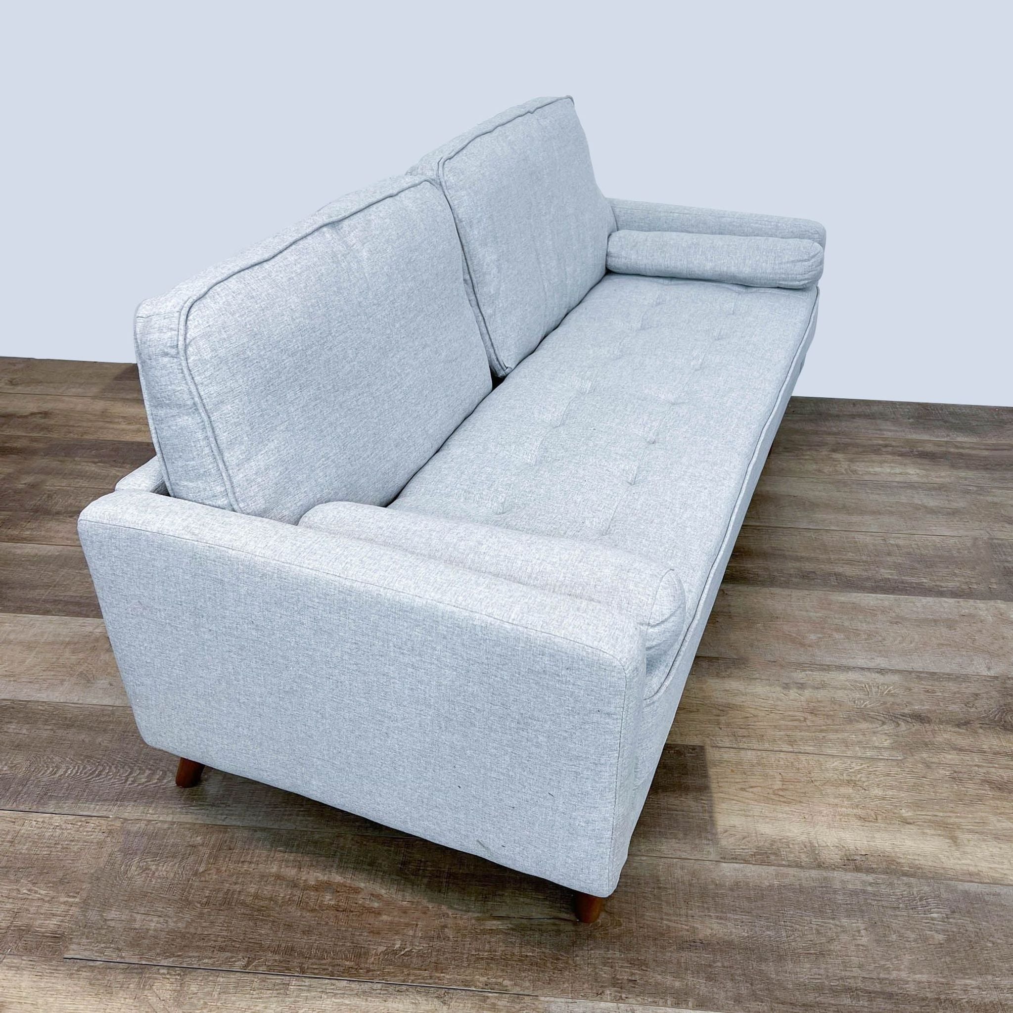 Reperch brand narrow arm loveseat with tufted bench seat, two bolster cushions, and wood finish feet against a wooden floor.