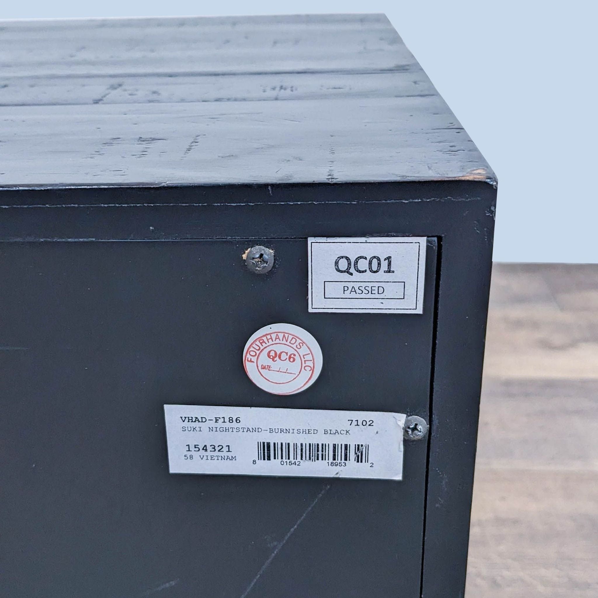 Back view of a Four Hands acacia wood end table showing quality control stickers and a barcode, indicating a burnished black finish and brass hardware.