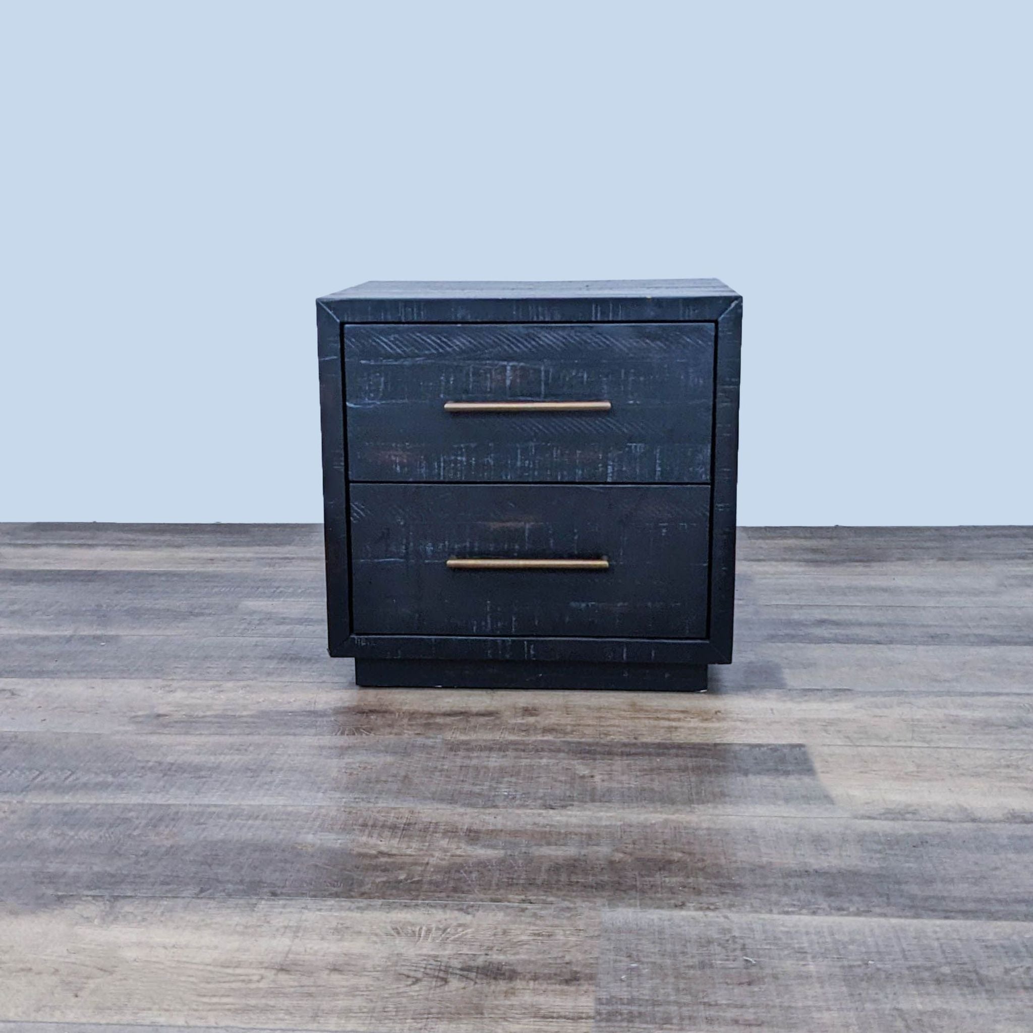 Four Hands brand acacia wood end table in a burnished black finish with two brass handles, on a wooden floor against a plain background.