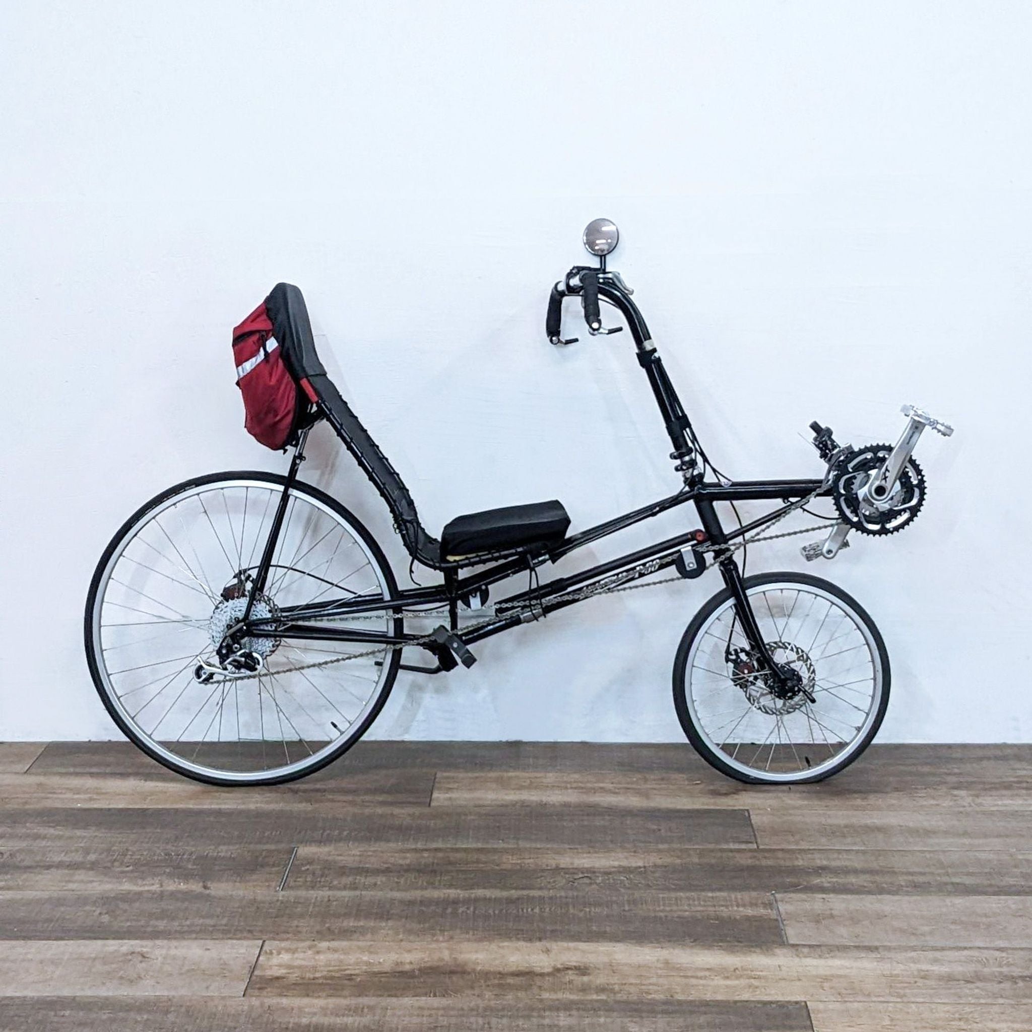 Alt text 1: Black Lightning P-38 recumbent bicycle with red bag mounted on rear, parked on a wood laminate floor against a white wall.