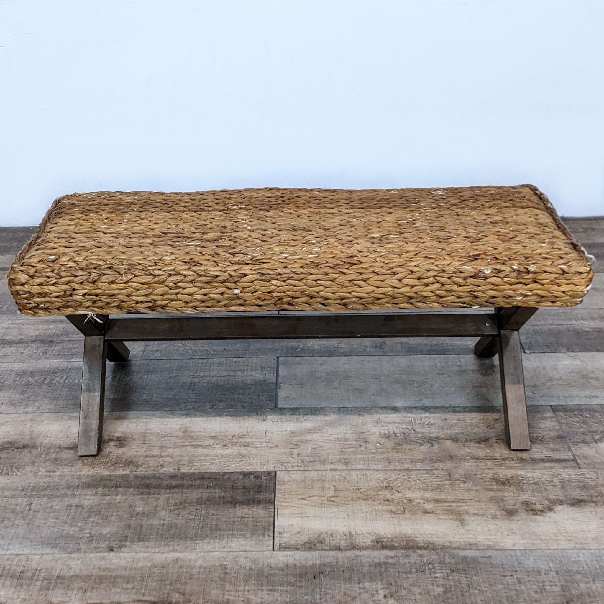 Alt text 1: A woven reed bench with a unique crisscross solid wood leg design by Reperch, sitting on a wooden floor.