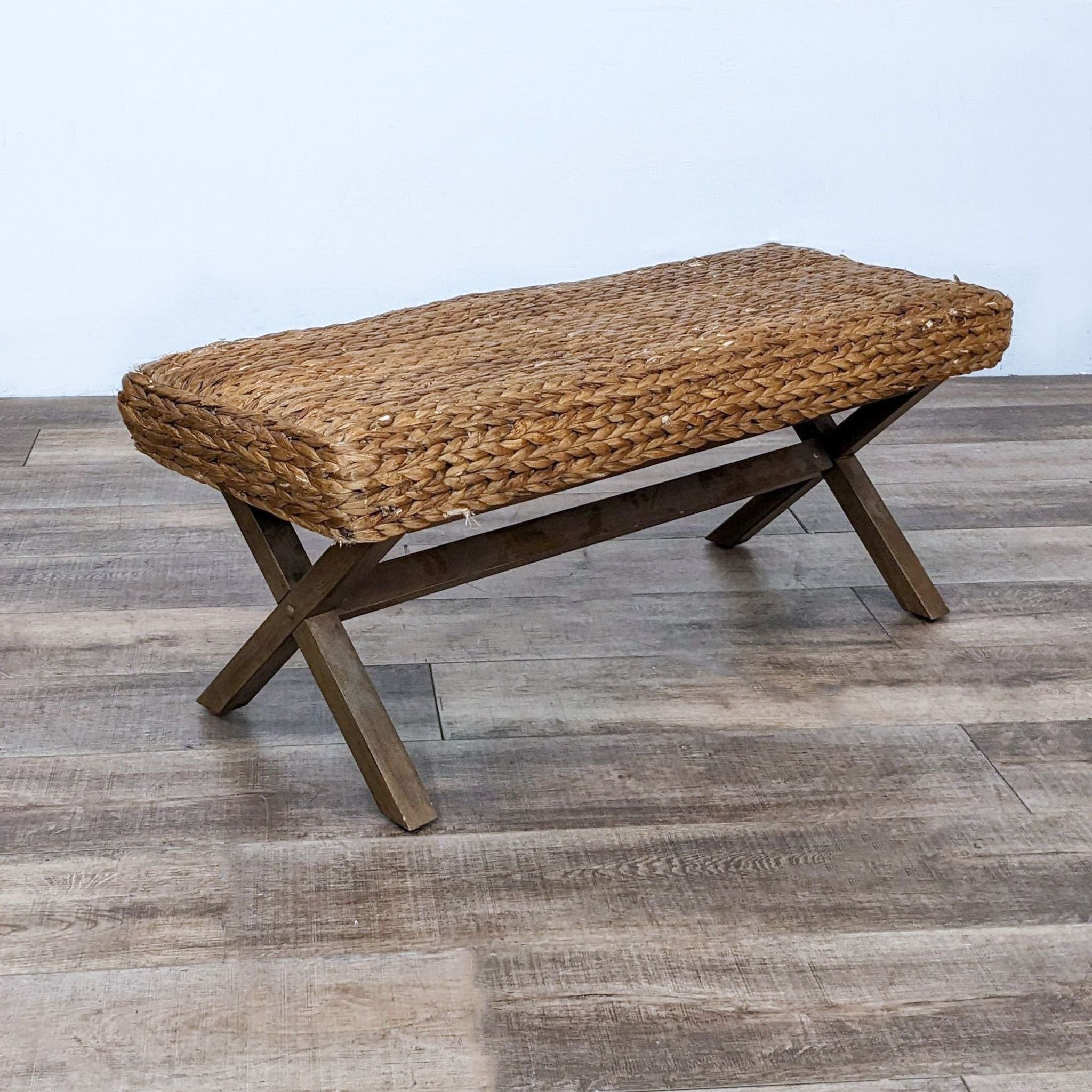 Alt text 2: Reperch's sturdy bench featuring a crisscross wooden base and woven reed top, placed against a neutral wall on a wood-patterned floor.