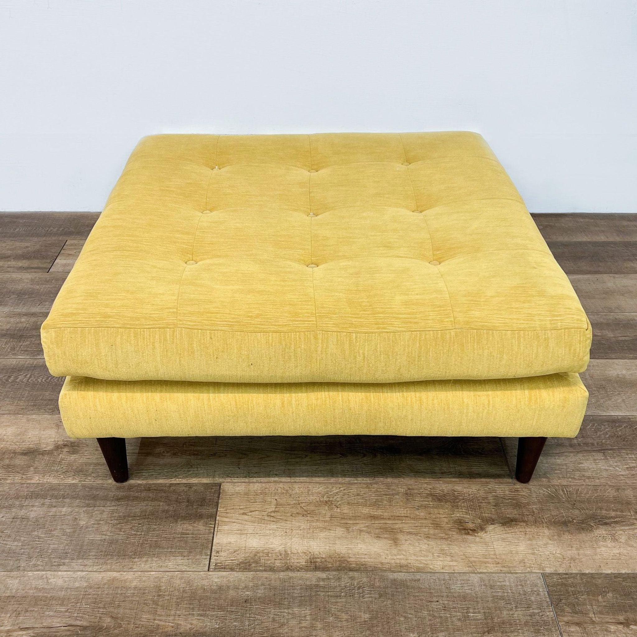 Square fabric tufted Joybird ottoman in yellow with wooden tapered legs, viewed from angle.