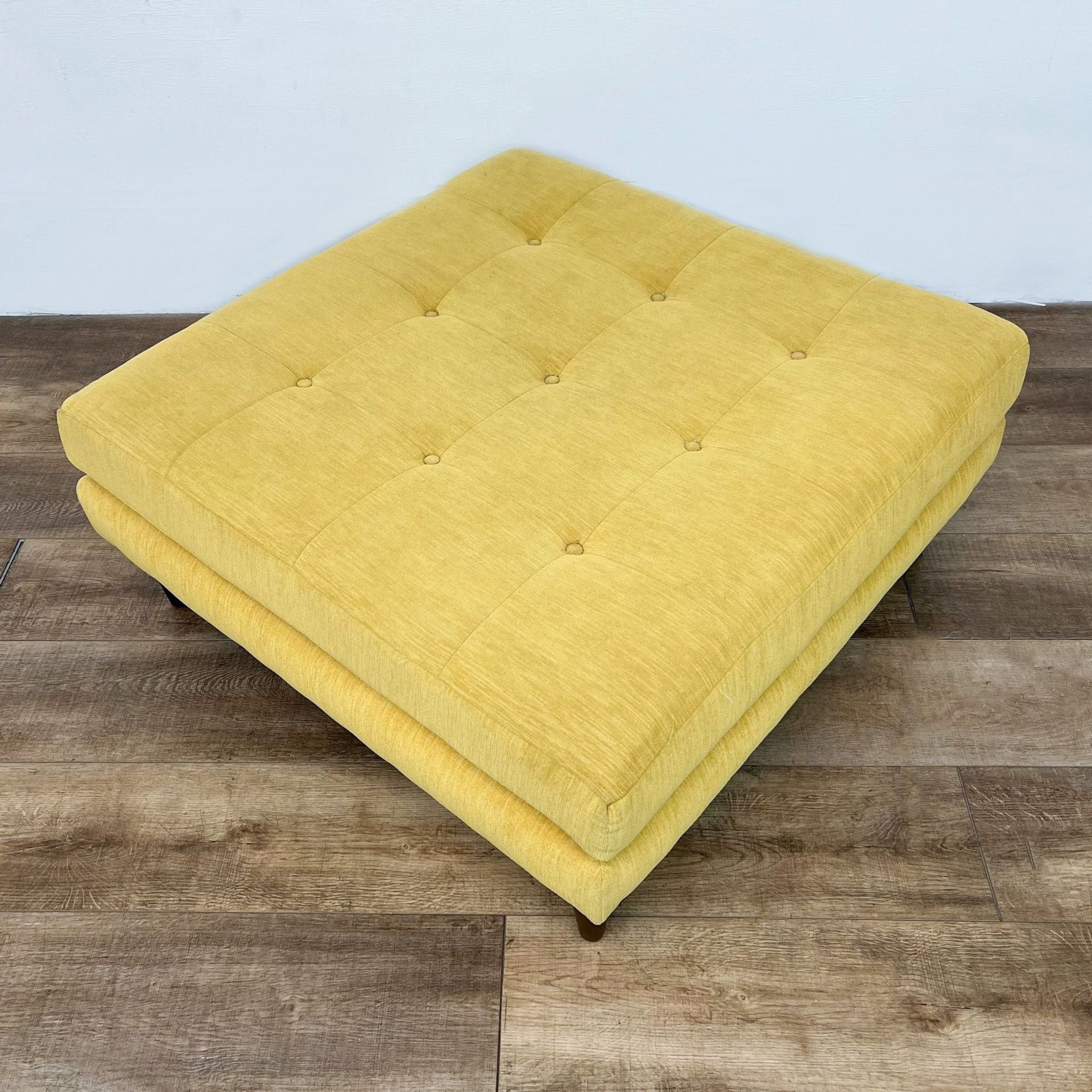 Yellow square Joybird ottoman with tufted fabric top and tapered wooden legs on a wood floor.