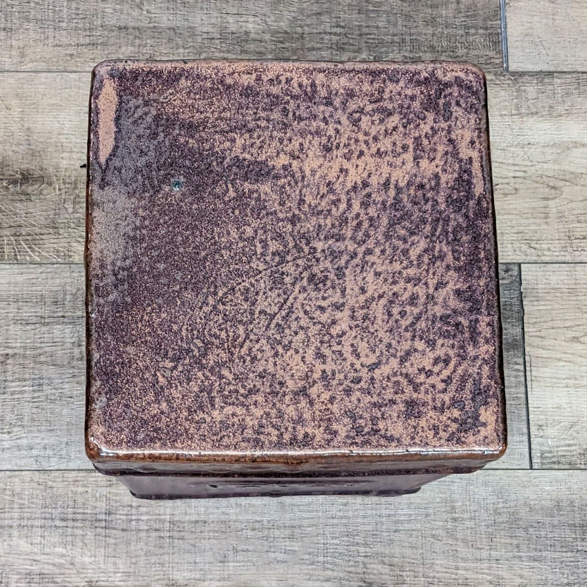 Top-down view of a square Reperch ceramic garden stool with a textured cinnabar glaze, on a wooden floor.
