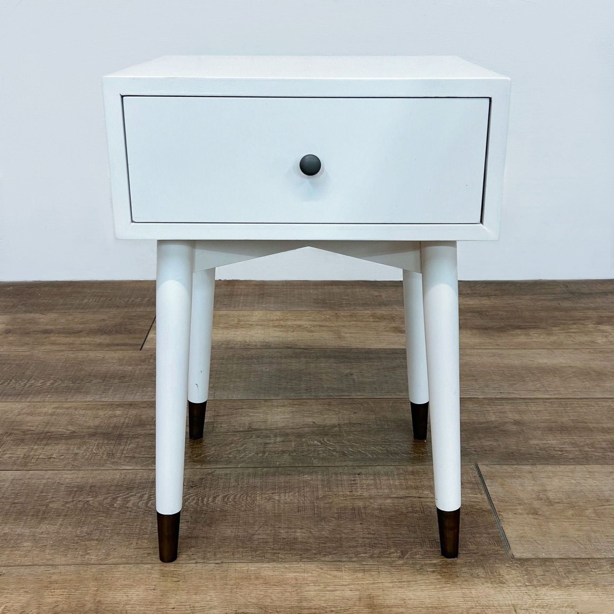 White Reperch end table with a single drawer and round knob, on a wood floor against a white wall.
