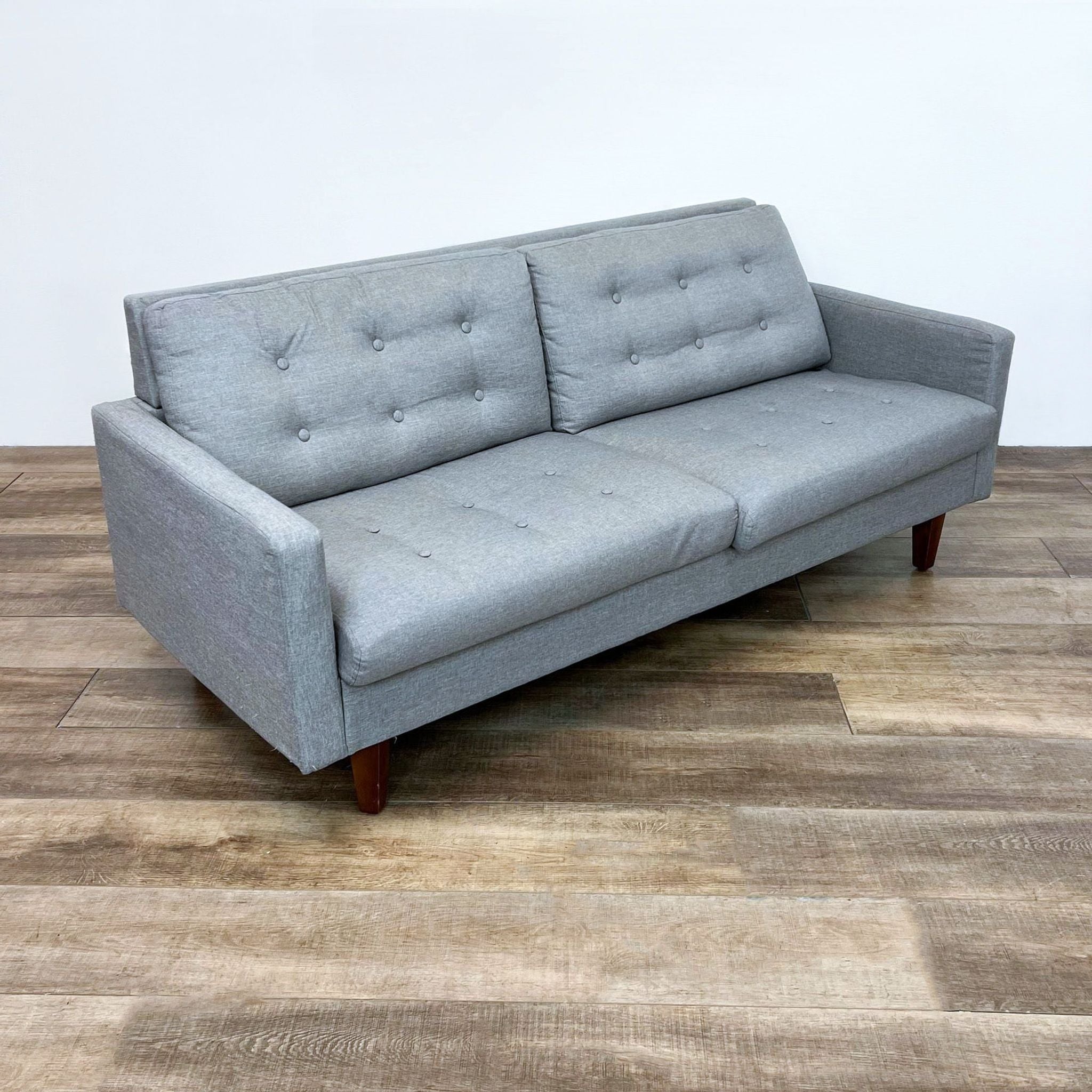 Gray upholstered three-seater Aeon Furniture sofa with button details and slanted wooden legs, placed on a wooden floor.