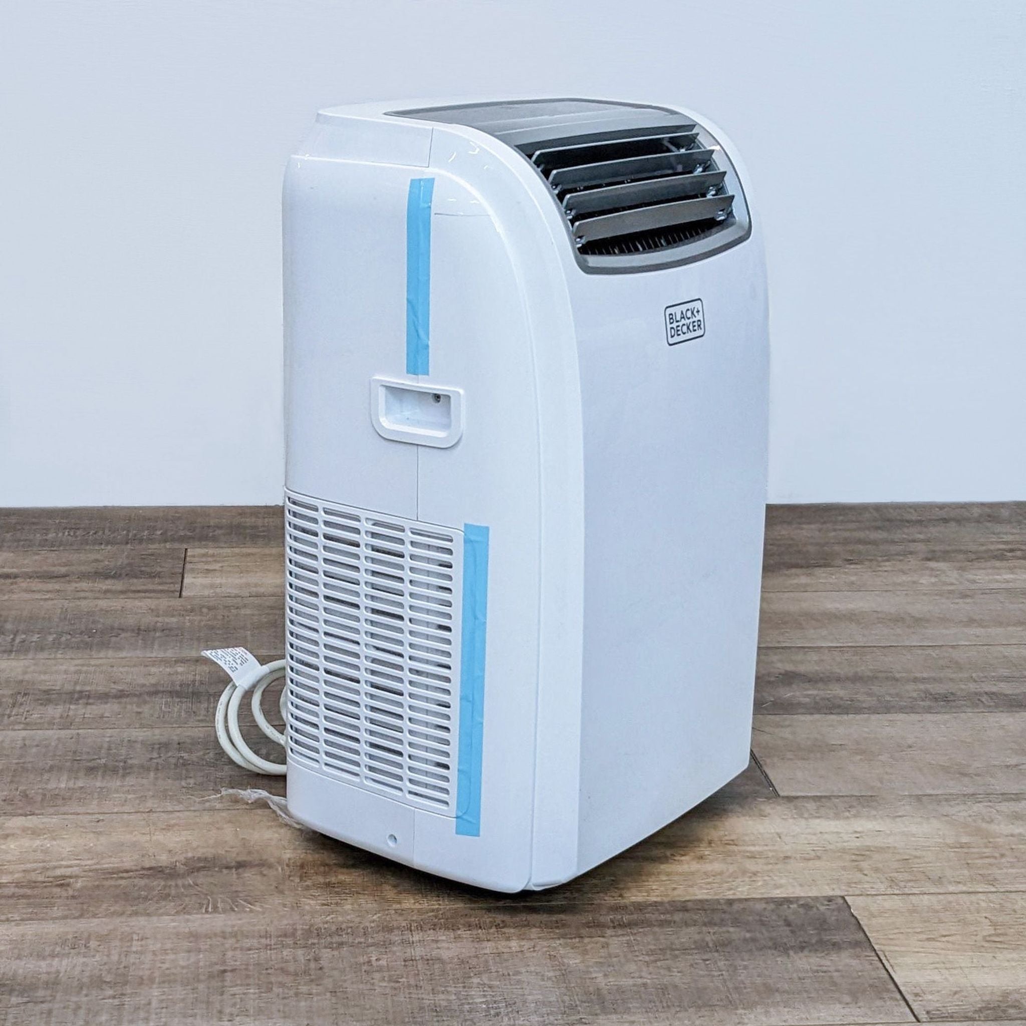 Image 2: Side angle of a Black + Decker portable air conditioner, showing vent and power cord on a wood-patterned floor.