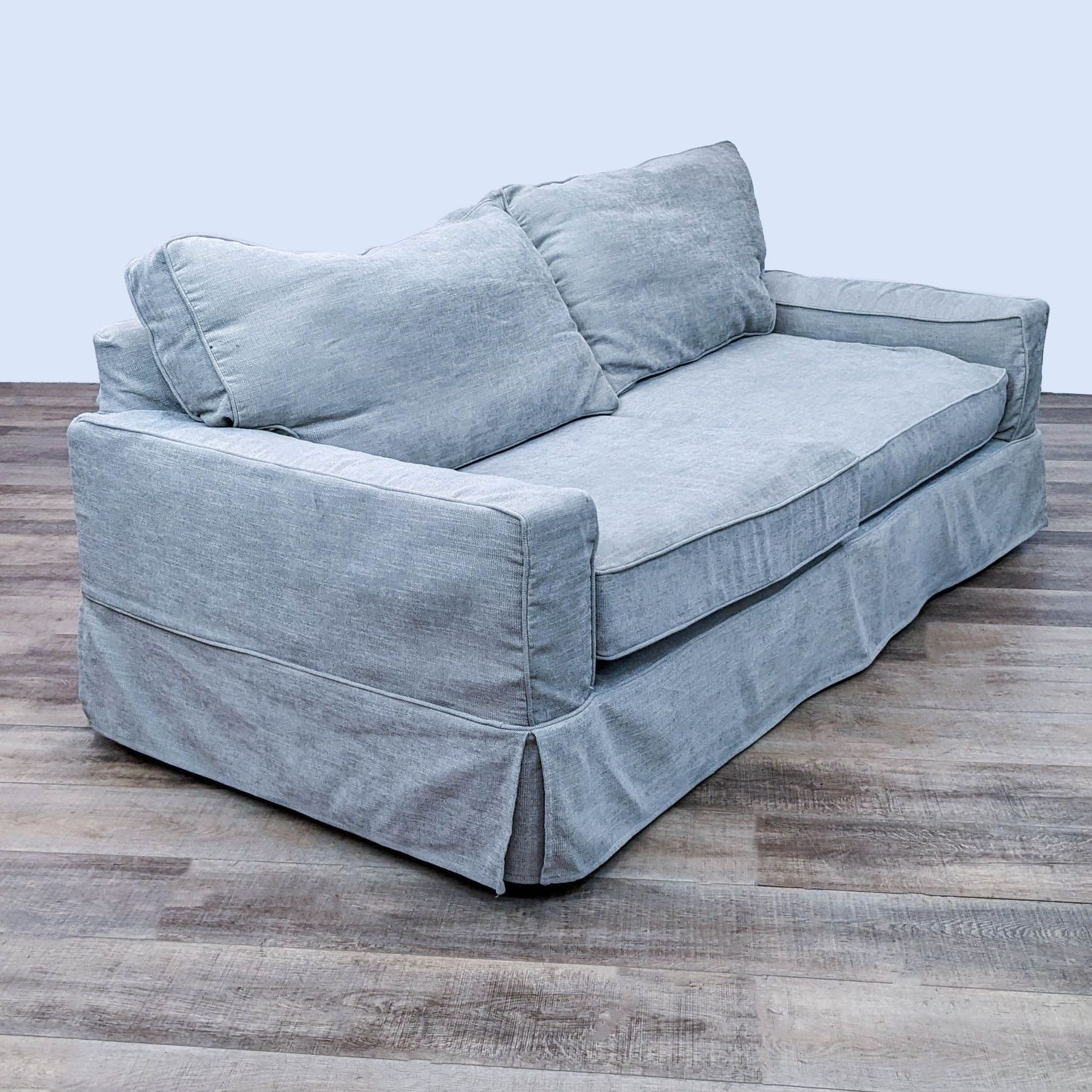 Angled view of a Pottery Barn gray fabric slipcover loveseat showcasing its design and track arms on a wooden surface.