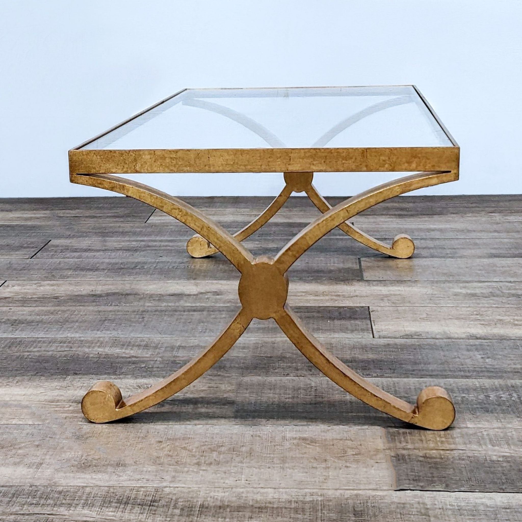 Alt text: David Iatesta coffee table with a glass top and a gilt-finished metal base, featuring a cross and scroll design on a wooden floor.