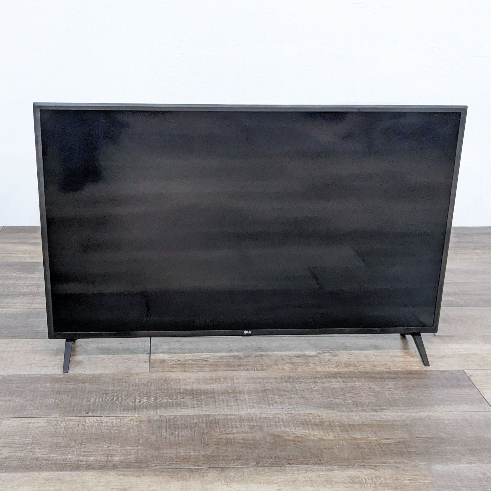 LG brand TV turned off, showing a black screen, on a wooden floor against a white wall.