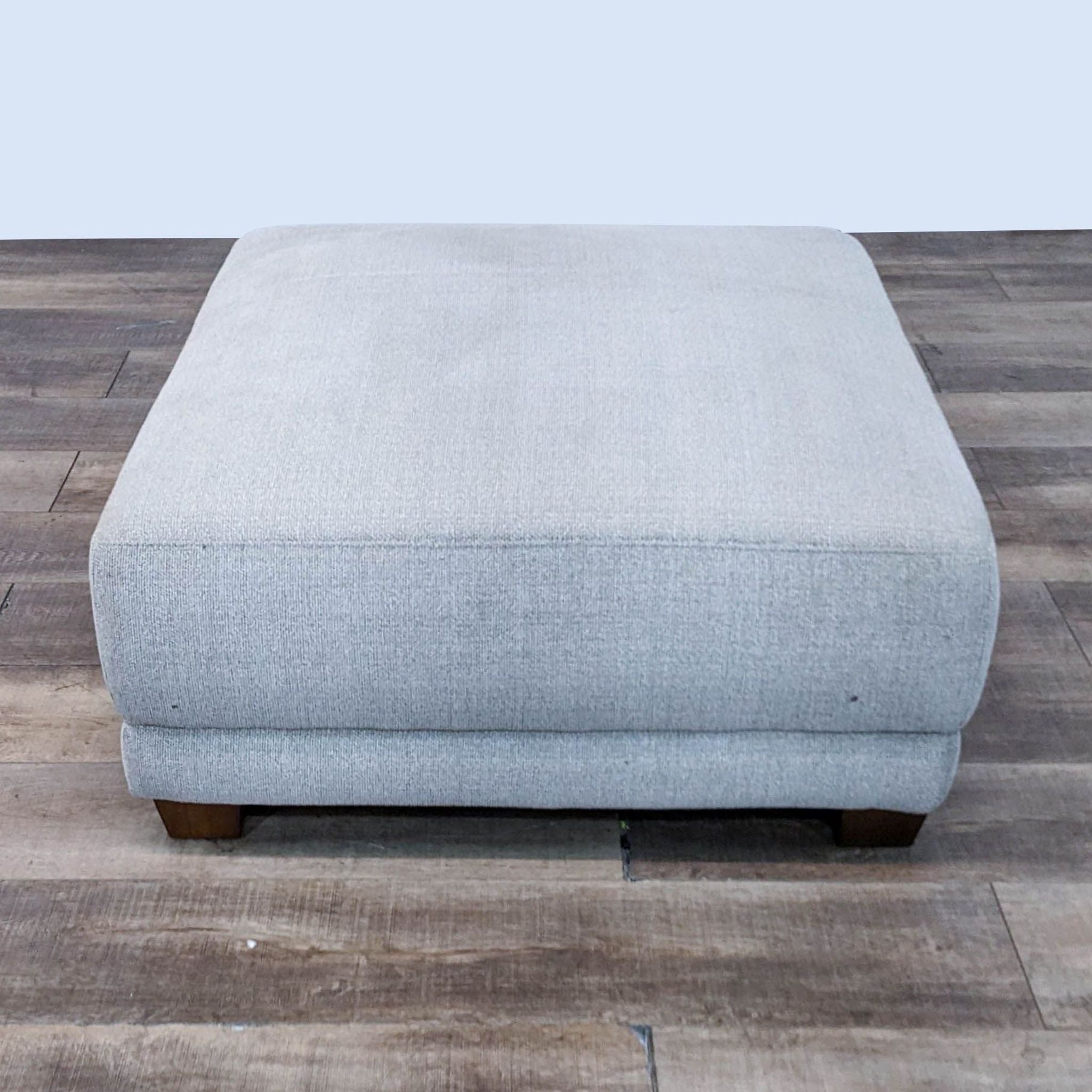 Alt text 1: A neutral-colored, square ottoman measuring 40 inches with dark-finished feet, by Costco, on a wooden floor.