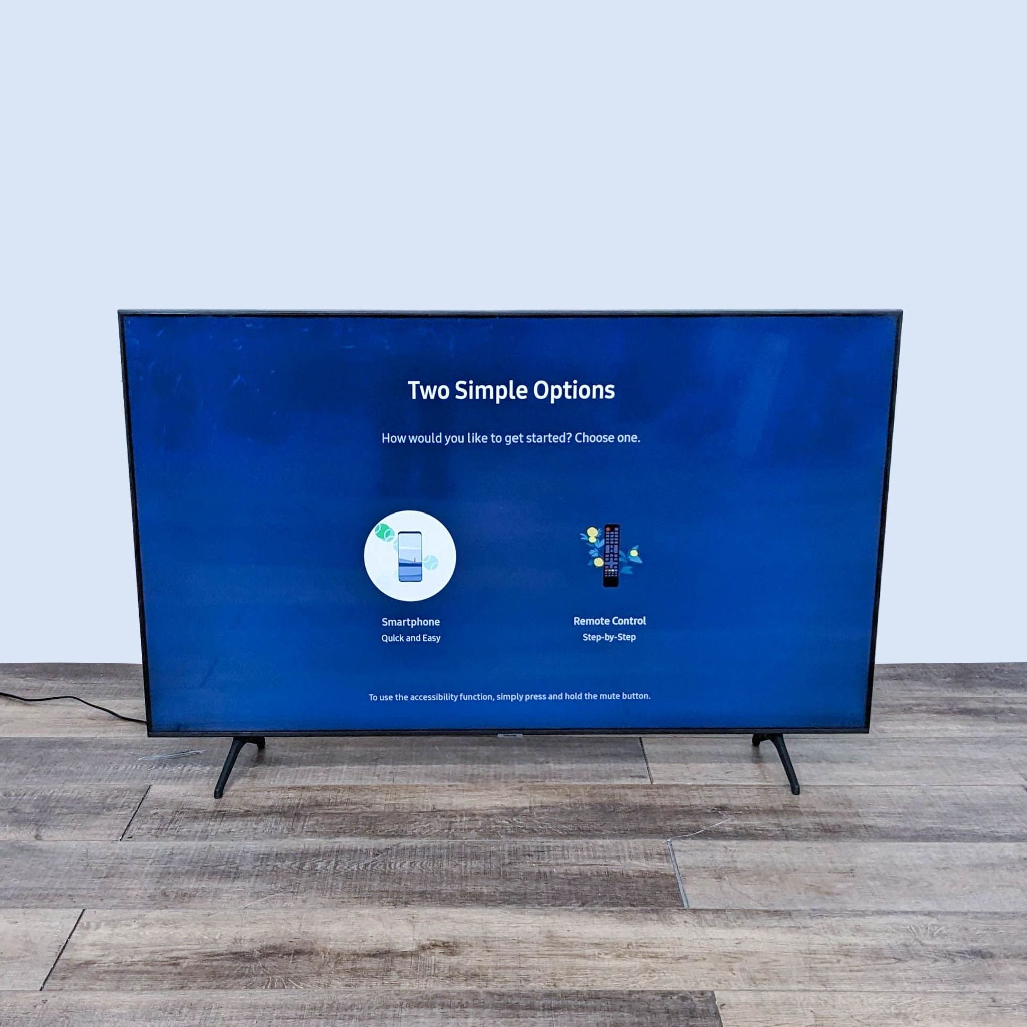 Modern Samsung flat screen TV on stand displaying a setup screen with options, against a plain wall and wood floor.