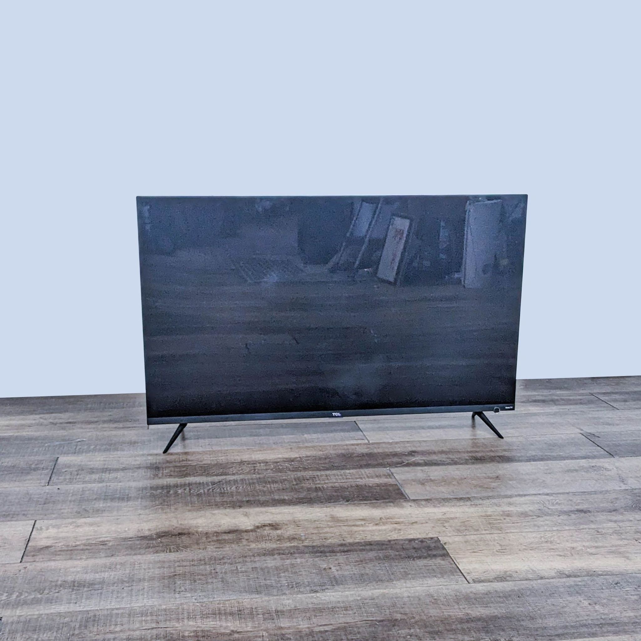 TCL television displayed in a frontal view, turned off, on a wooden floor against a grey wall.