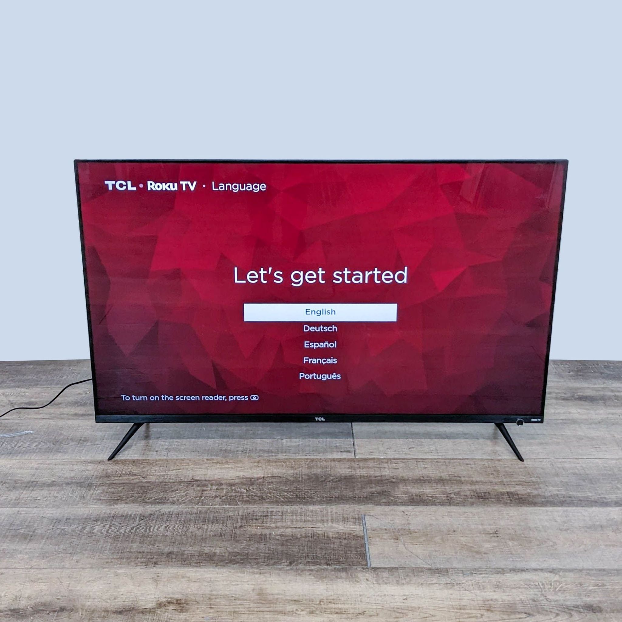 TCL Roku TV on startup screen with language selection options, standing on a wooden floor against a white background.