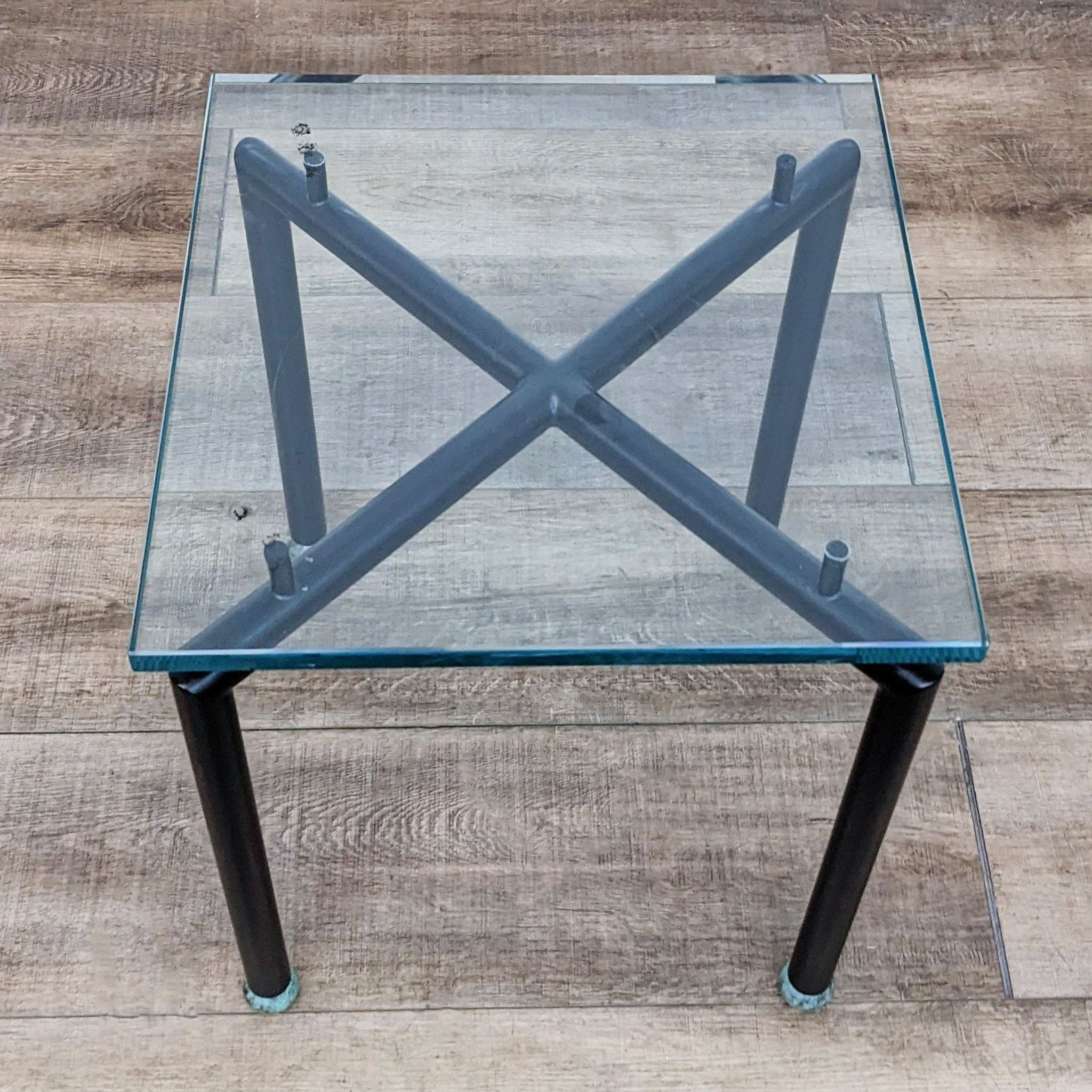 Glass top console table by Reperch with a metal X-shaped base structure and blue edges, on wooden flooring.