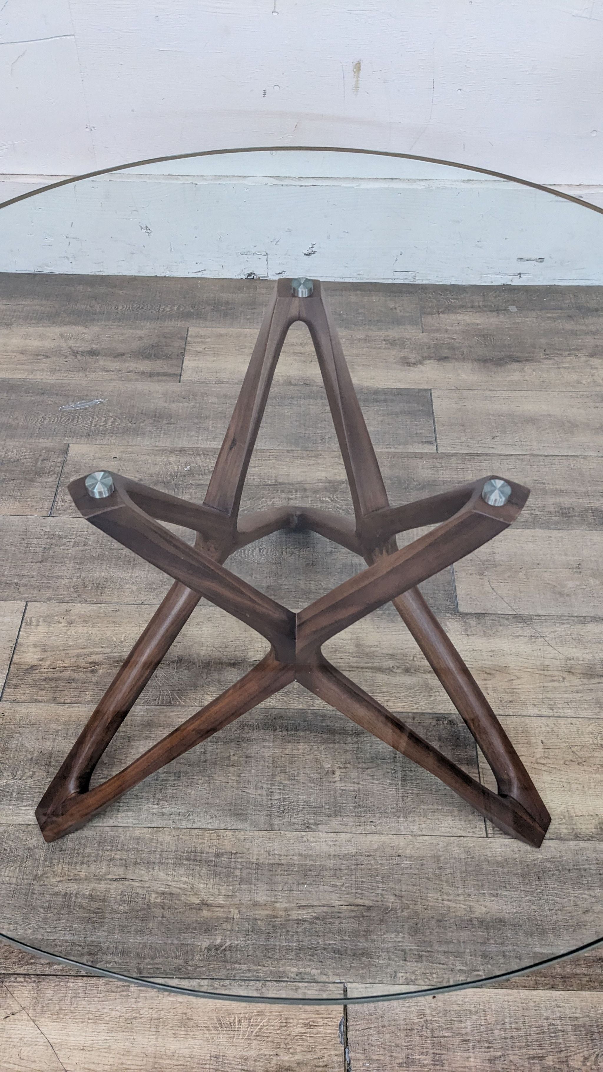 Scandinavian Design dining table with circular glass top and a wooden cross base on a wooden floor.