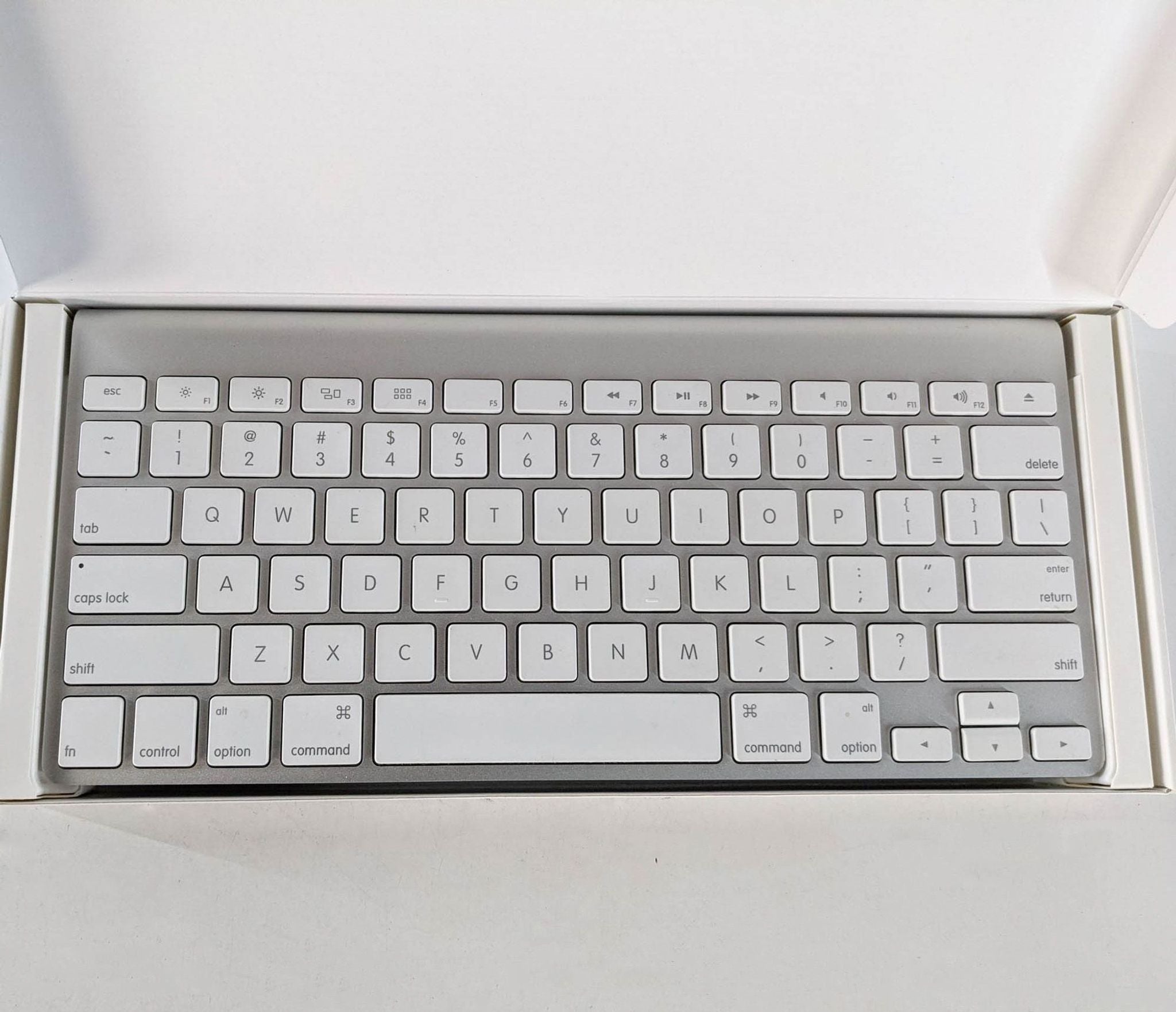 Alt text 2: Apple wireless keyboard in open box, viewed from above, highlighting the sleek layout and white keys against a white background.