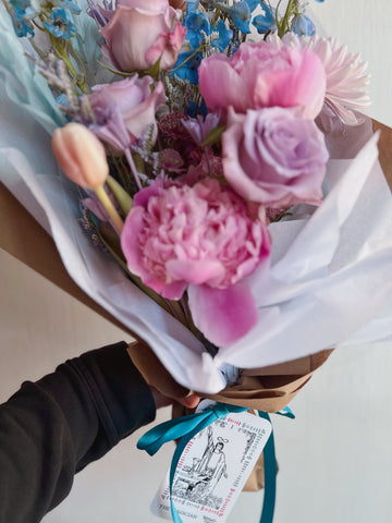 A wrapped fresh flower bouquet with soft lavender, pink, and blue flowers and a The Magician tarot card tied to it