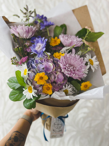 A fresh flower arrangement wrapped in paper, featuring lavender mums and daisies, with a Wheel of Fortune tarot card tied around it