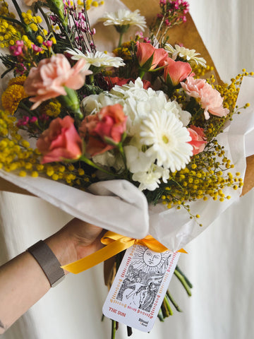 A fresh flower bouquet with a The Sun tarot card tied to it with ribbon