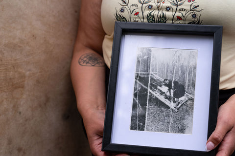 hands hold a historic black and white framed photo of an Indigenous grandmother scraping a moose hide on a frame outdoors.