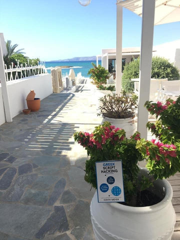A photo of a balcony in Greece with a bright blue sky, and a copy of Greek Script Hacking hiding in a flower pot