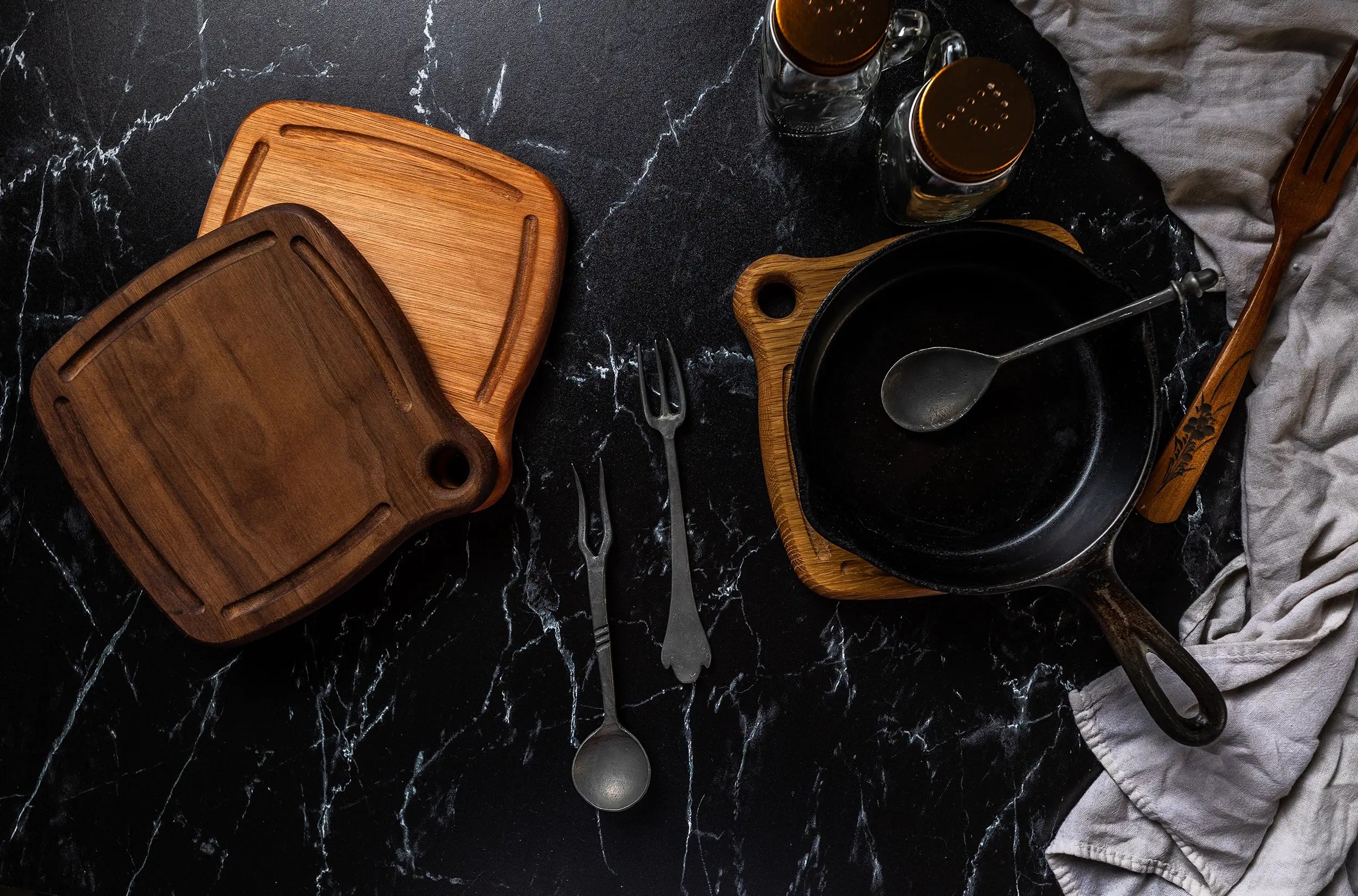 A black cast iron skillet with a metal spoon inside rests on a wooden trivet, accompanied by a pair of Trivets, salt and pepper shakers, and antique-style serving utensils on a black marble countertop. A linen cloth is draped in the corner.