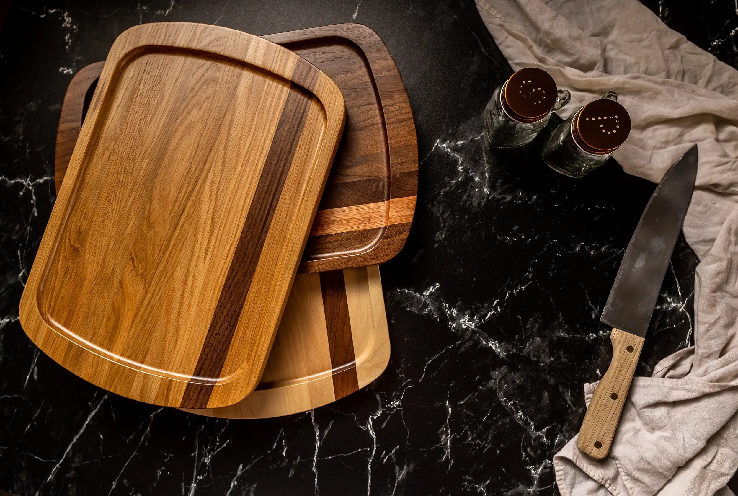 Stylish serving trays with alternating wood tones sit fanned out on a glossy black surface, alongside a wooden-handled knife and two glass spice jars.
