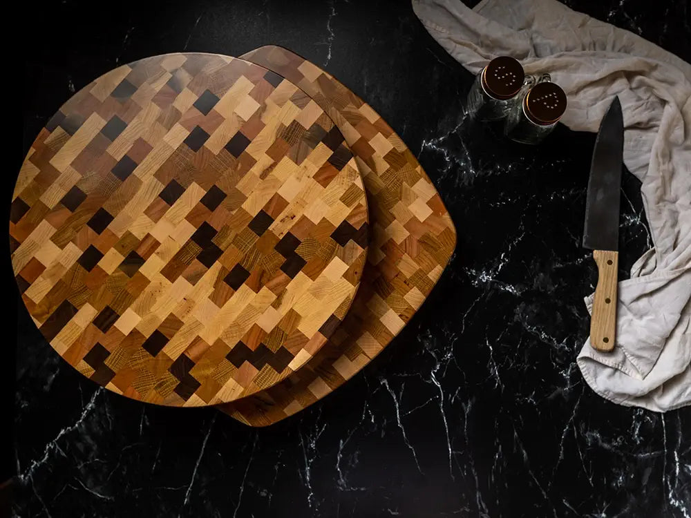Two wooden cutting boards with a pixelated pattern of various wood tones are stacked on a black marble countertop beside a chef’s knife, two spice shakers, and a beige cloth.