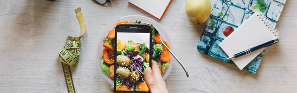 a hand holding a phone, taking a photo of his meal. A measuring ribbon to the left of the picture.