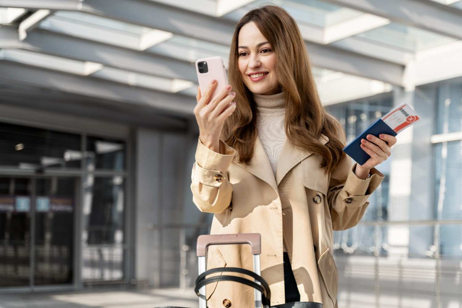 Smiling woman outside airport using smartphone with eSIM, holding passport and boarding pass, suitcase in foreground.