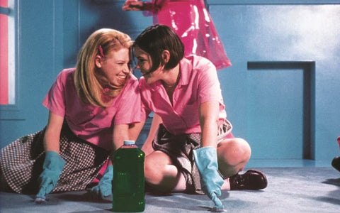 Two girls kneeling on the ground with large rubber gloves on, smiling at each other as they clean the floor.
