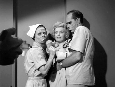 Two nurses with disfigured faces hold a shocked woman back.