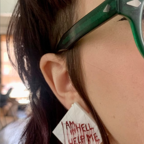 A close up photo of handmade earrings inspired by Hellraiser 2, made to look like the bathroom wall in the film, with bloody txt that reads "Help Me I Am In Hell"