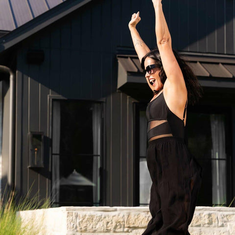 Woman in black maxi dress and sunglasses cheering with her arms in the air