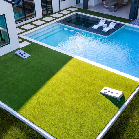 Backyard with large rectangle of grass next to rectangle pool with ledge. Pool ledge has two white Ledge Lounger Signature Chaises, and grass and a white cornhole game