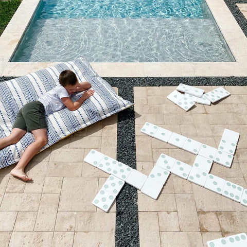 Child laying on Ledge Lounger Laze Pillow and looking at dominoes laid out on patio in front of pool