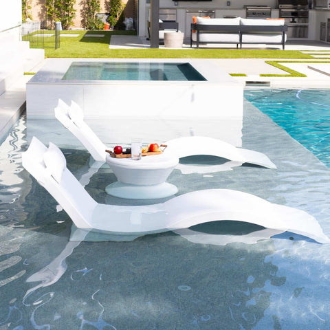 A pair of luxurious pool loungers on the tanning ledge of a backyard pool, with a charcuterie board a top an in pool side table.