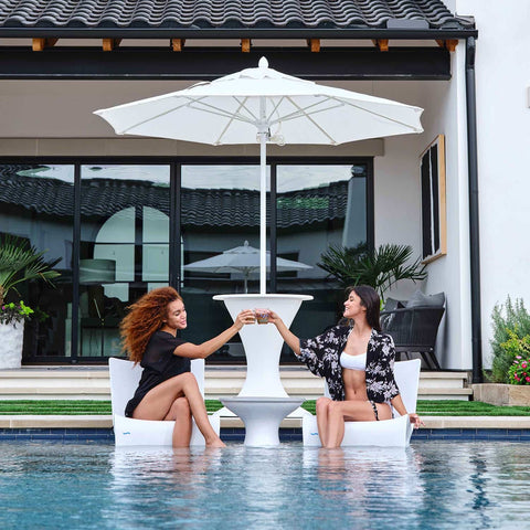 A pair of girl friends enjoying their in pool chairs while chatting over drinks.