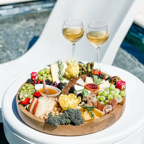 Charcuterie board with two glasses of white wine on in-pool side table, with Ledge Lounger in-pool chaise behind it