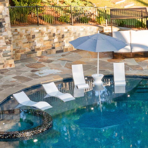 A backyard pool's tanning ledge fully set up with in pool chaises, chairs, side tables, and an umbrella.