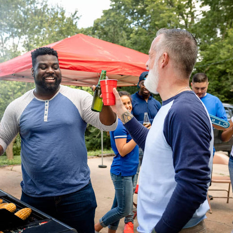 Two men "cheers" drinks over the grill at a tailgate