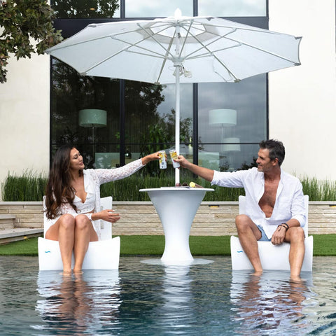 Two adults sitting in Ledge Loungers in-pool chairs toast with tall cans in front of an in-pool tall side table and umbrella