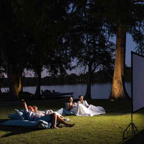 Four people lounging on two Ledge Lounger Laze Pillows on grass in front of a projection screen in a backyard at night